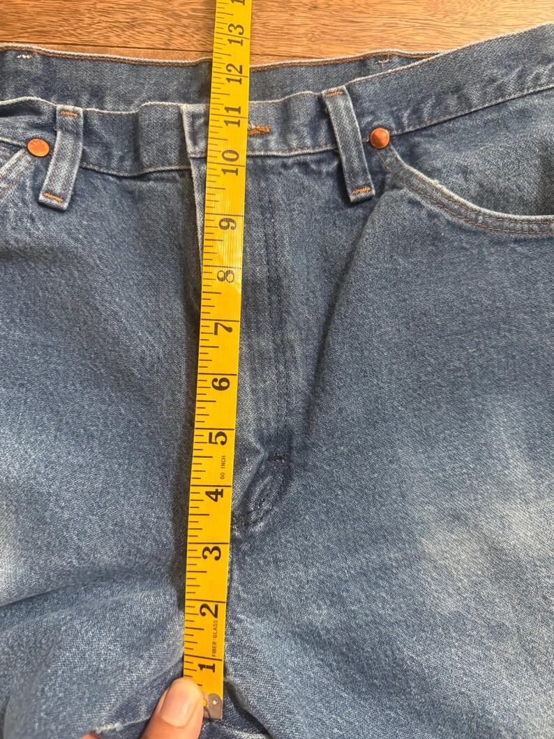 May include: Close-up of blue denim jeans with a yellow measuring tape. The tape shows a measurement of approximately 13 inches. The jeans have copper rivets and a classic five-pocket design.