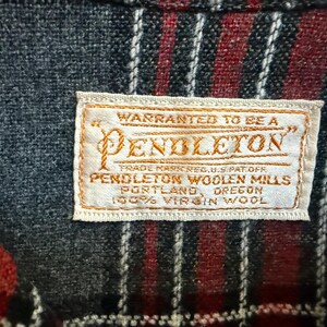 Może przedstawiać: Zbliżenie metki Pendleton wszytej w tkaninę w kratę. Etykieta ze złotymi literami głosi "WARRANTED TO BE A PENDLETON" i inne szczeg&oacute;ły. Tkanina ma wz&oacute;r czerwonych i czarnych pask&oacute;w.