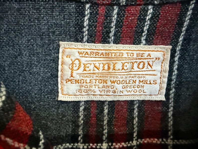 Może przedstawiać: Zbliżenie metki Pendleton wszytej w tkaninę w kratę. Etykieta ze złotymi literami głosi "WARRANTED TO BE A PENDLETON" i inne szczeg&oacute;ły. Tkanina ma wz&oacute;r czerwonych i czarnych pask&oacute;w.