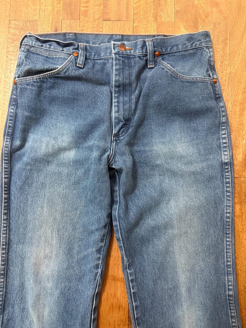 May include: Blue denim jeans with a classic five-pocket design. The jeans have a faded wash and copper-colored rivets. The waist features a button closure and belt loops. The jeans are made of durable denim.