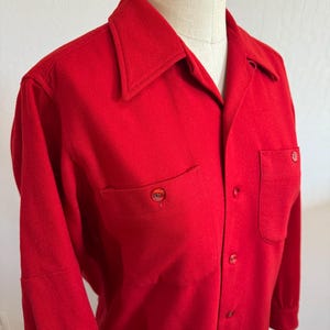 以下が含まれることがあります： 鮮やかな赤色のボタンダウンシャツ。胸に2つのポケットと尖った襟が付いています。長袖シャツにはお揃いの赤いボタンが付いており、マネキンに着せられています。生地は中程度の厚さに見えます。