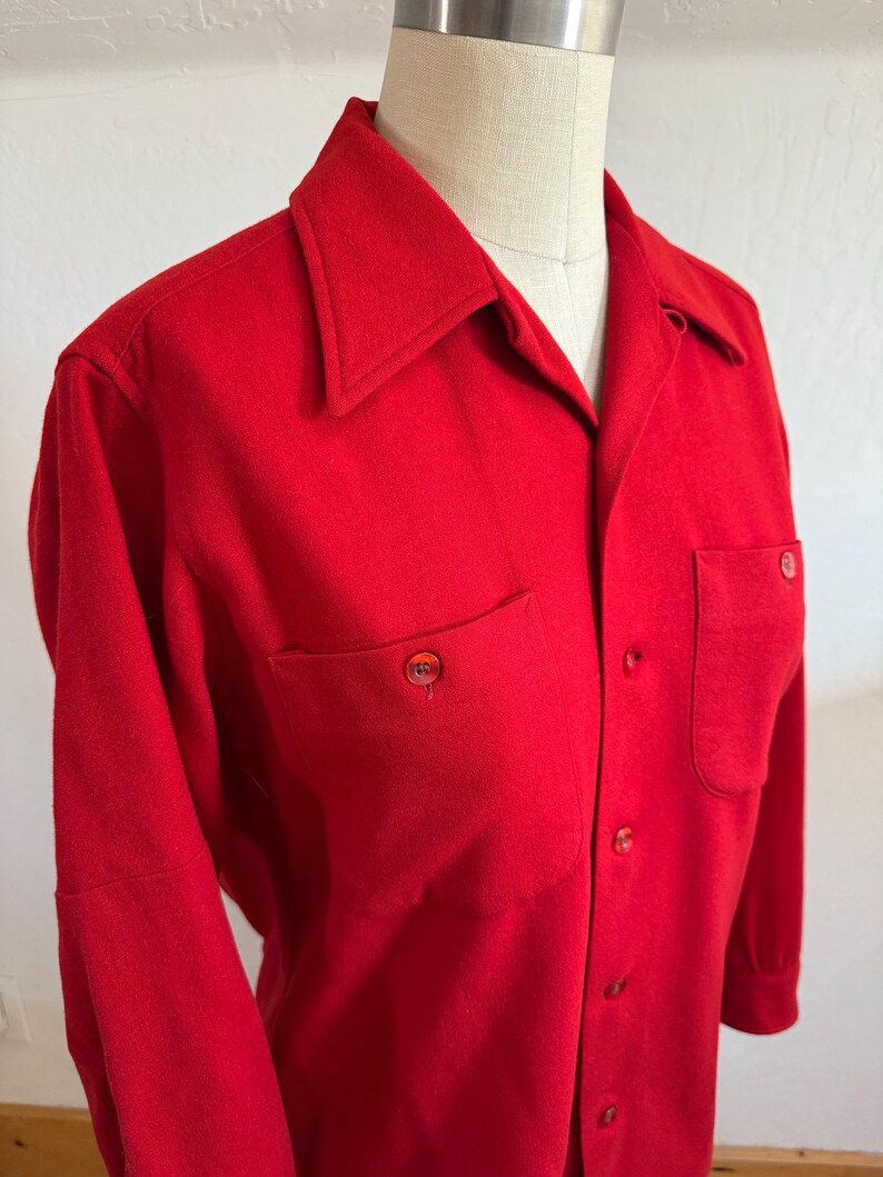 以下が含まれることがあります： 鮮やかな赤色のボタンダウンシャツ。胸に2つのポケットと尖った襟が付いています。長袖シャツにはお揃いの赤いボタンが付いており、マネキンに着せられています。生地は中程度の厚さに見えます。