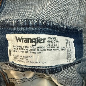 K&ouml;nnte beinhalten: Nahaufnahme eines Wrangler Jeansetiketts. Das Etikett ist wei&szlig; mit schwarzem Text, einschlie&szlig;lich Waschanleitung und Gr&ouml;&szlig;e 33 x 32. Die Jeans ist aus blauem Denim, mit sichtbaren N&auml;hten und einem Knopf.