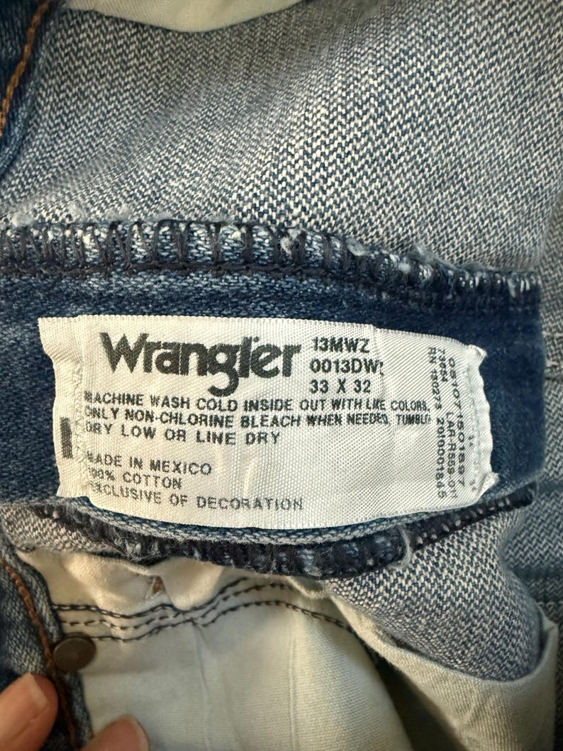 K&ouml;nnte beinhalten: Nahaufnahme eines Wrangler Jeansetiketts. Das Etikett ist wei&szlig; mit schwarzem Text, einschlie&szlig;lich Waschanleitung und Gr&ouml;&szlig;e 33 x 32. Die Jeans ist aus blauem Denim, mit sichtbaren N&auml;hten und einem Knopf.