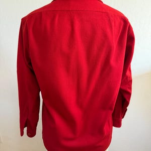 以下が含まれることがあります： 鮮やかな赤色の長袖シャツ。ボタンダウンの襟とボタン付きの袖口が特徴です。マネキンに着せられており、衣服の背面が示されています。生地は中程度の厚さに見えます。