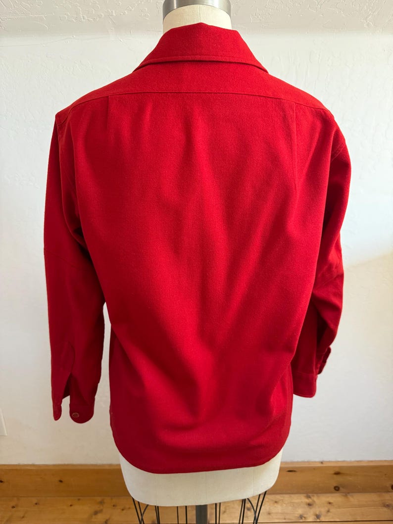 以下が含まれることがあります： 鮮やかな赤色の長袖シャツ。ボタンダウンの襟とボタン付きの袖口が特徴です。マネキンに着せられており、衣服の背面が示されています。生地は中程度の厚さに見えます。