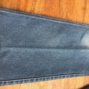Pu&ograve; includere: Primo piano di jeans blu con un metro a nastro giallo. Il nastro mostra che la larghezza della gamba dei jeans &egrave; di circa 20 cm. I jeans sono su una superficie di legno.