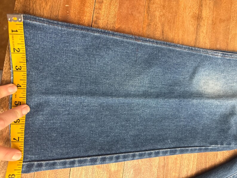 Pu&ograve; includere: Primo piano di jeans blu con un metro a nastro giallo. Il nastro mostra che la larghezza della gamba dei jeans &egrave; di circa 20 cm. I jeans sono su una superficie di legno.