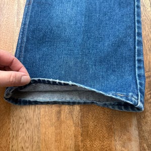 Pu&ograve; includere: Primo piano di jeans di denim blu. I jeans hanno un classico design a cinque tasche e una gamba dritta. Il denim &egrave; un lavaggio medio con qualche sbiadimento e usura. L'orlo &egrave; ripiegato, rivelando l'interno dei jeans.