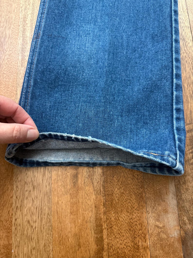 Pu&ograve; includere: Primo piano di jeans di denim blu. I jeans hanno un classico design a cinque tasche e una gamba dritta. Il denim &egrave; un lavaggio medio con qualche sbiadimento e usura. L'orlo &egrave; ripiegato, rivelando l'interno dei jeans.