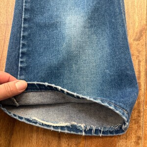 Pu&ograve; includere: Primo piano di un paio di jeans di denim blu. L'orlo &egrave; sfilacciato e il tessuto mostra segni di usura. I jeans sono di un lavaggio medio, con aree pi&ugrave; chiare dovute all'uso.