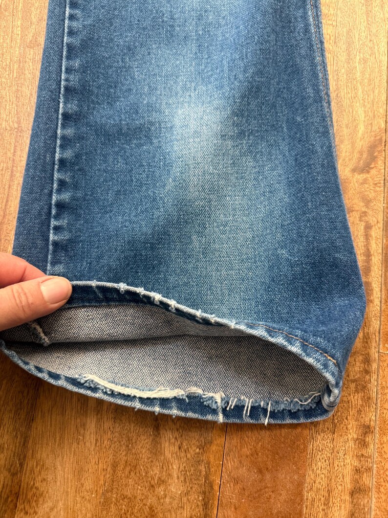 Pu&ograve; includere: Primo piano di un paio di jeans di denim blu. L'orlo &egrave; sfilacciato e il tessuto mostra segni di usura. I jeans sono di un lavaggio medio, con aree pi&ugrave; chiare dovute all'uso.
