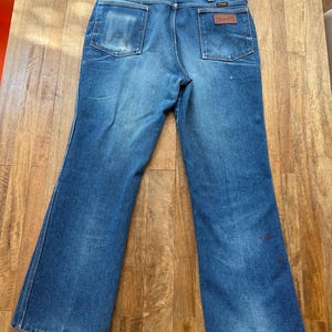 Pu&ograve; includere: Jeans Wrangler vintage in denim blu con gamba svasata. I jeans hanno due tasche posteriori, una con toppa in pelle marrone. Il denim mostra segni di usura e sbiadimento, con qualche piccola macchia.