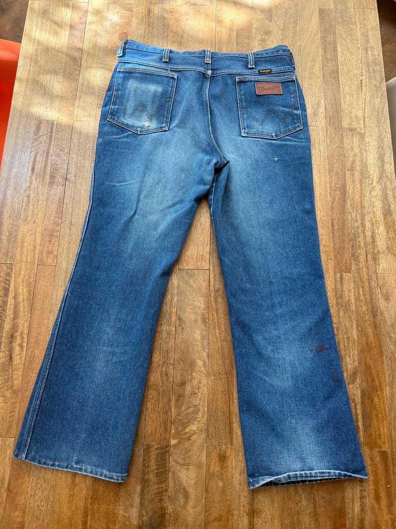Pu&ograve; includere: Jeans Wrangler vintage in denim blu con gamba svasata. I jeans hanno due tasche posteriori, una con toppa in pelle marrone. Il denim mostra segni di usura e sbiadimento, con qualche piccola macchia.