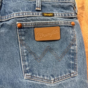 May include: Close-up of blue denim jeans with a back pocket. The pocket features a brown leather patch with the "Wrangler" logo. The jeans have copper-colored rivets and a small "Wrangler" label above the pocket.