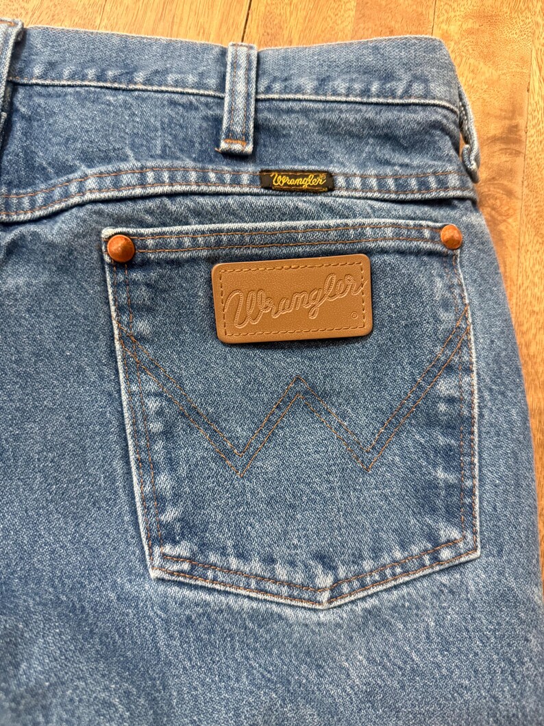 May include: Close-up of blue denim jeans with a back pocket. The pocket features a brown leather patch with the "Wrangler" logo. The jeans have copper-colored rivets and a small "Wrangler" label above the pocket.