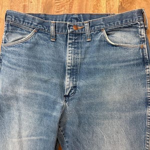 May include: Close-up of light blue denim jeans. The jeans have a classic five-pocket design, a button and zipper closure, and belt loops. The denim has a faded appearance, with subtle wear marks.