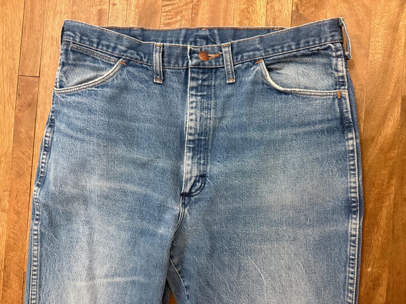 May include: Close-up of light blue denim jeans. The jeans have a classic five-pocket design, a button and zipper closure, and belt loops. The denim has a faded appearance, with subtle wear marks.