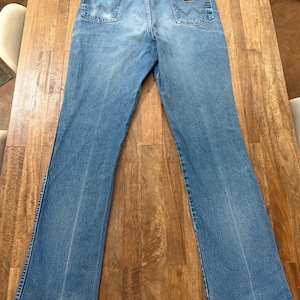 May include: Light blue denim jeans with a classic five-pocket design. The jeans feature a brown leather patch on the back pocket and a faded wash. The jeans are laid flat on a wooden surface.