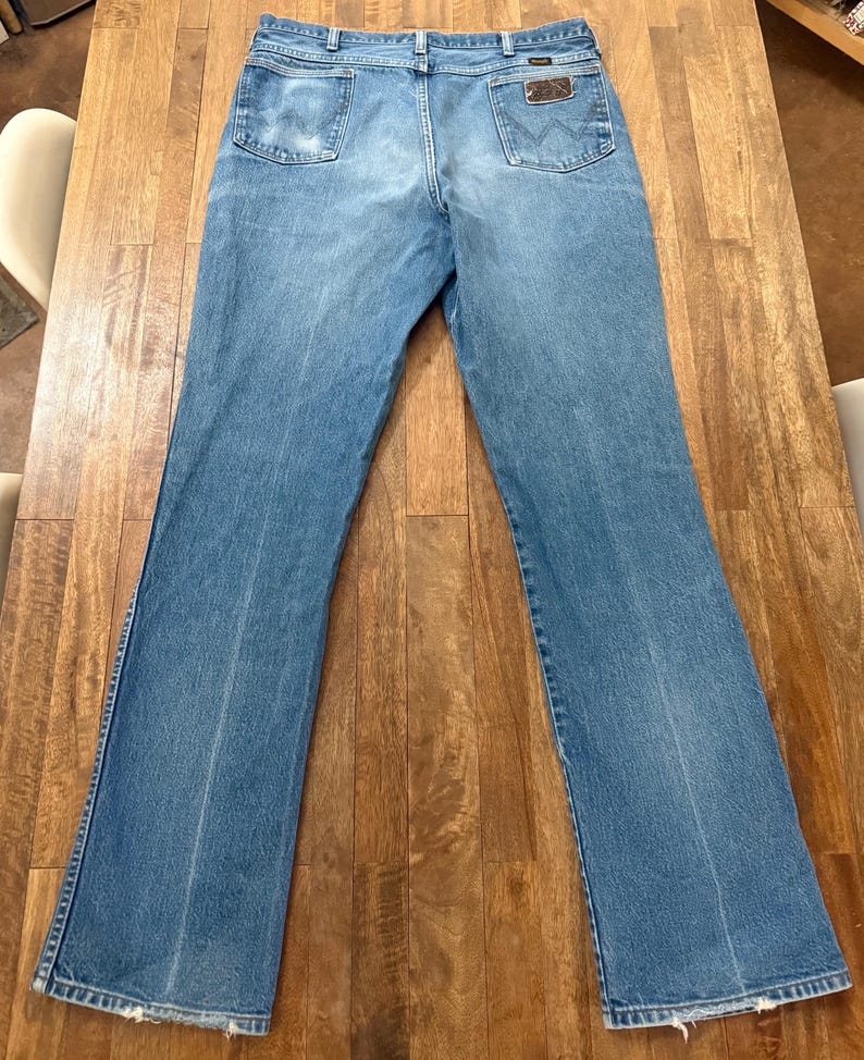May include: Light blue denim jeans with a classic five-pocket design. The jeans feature a brown leather patch on the back pocket and a faded wash. The jeans are laid flat on a wooden surface.