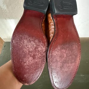 May include: Pair of brown leather cowboy boots, showing the soles. The soles are a deep burgundy color, with visible wear and stitching. The heels are black rubber, and the boots appear to be well-worn.