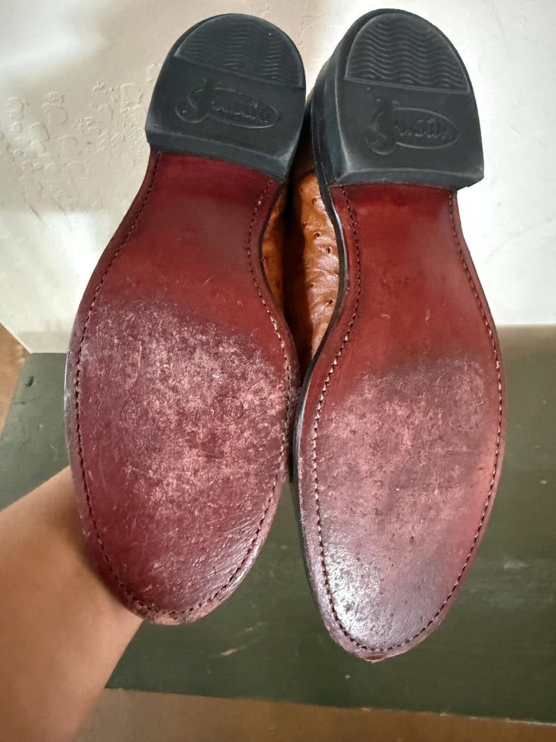 May include: Pair of brown leather cowboy boots, showing the soles. The soles are a deep burgundy color, with visible wear and stitching. The heels are black rubber, and the boots appear to be well-worn.