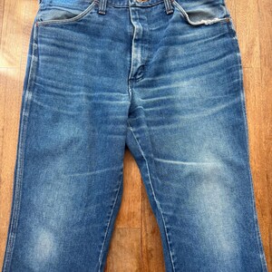 Pu&ograve; includere: Un paio di jeans vintage in denim blu. I jeans hanno un classico design a cinque tasche, una chiusura con bottone e cerniera e un lavaggio sbiadito. Il denim mostra segni di usura, con qualche sbiaditura e usura. I jeans sono appoggiati su una superficie di legno.