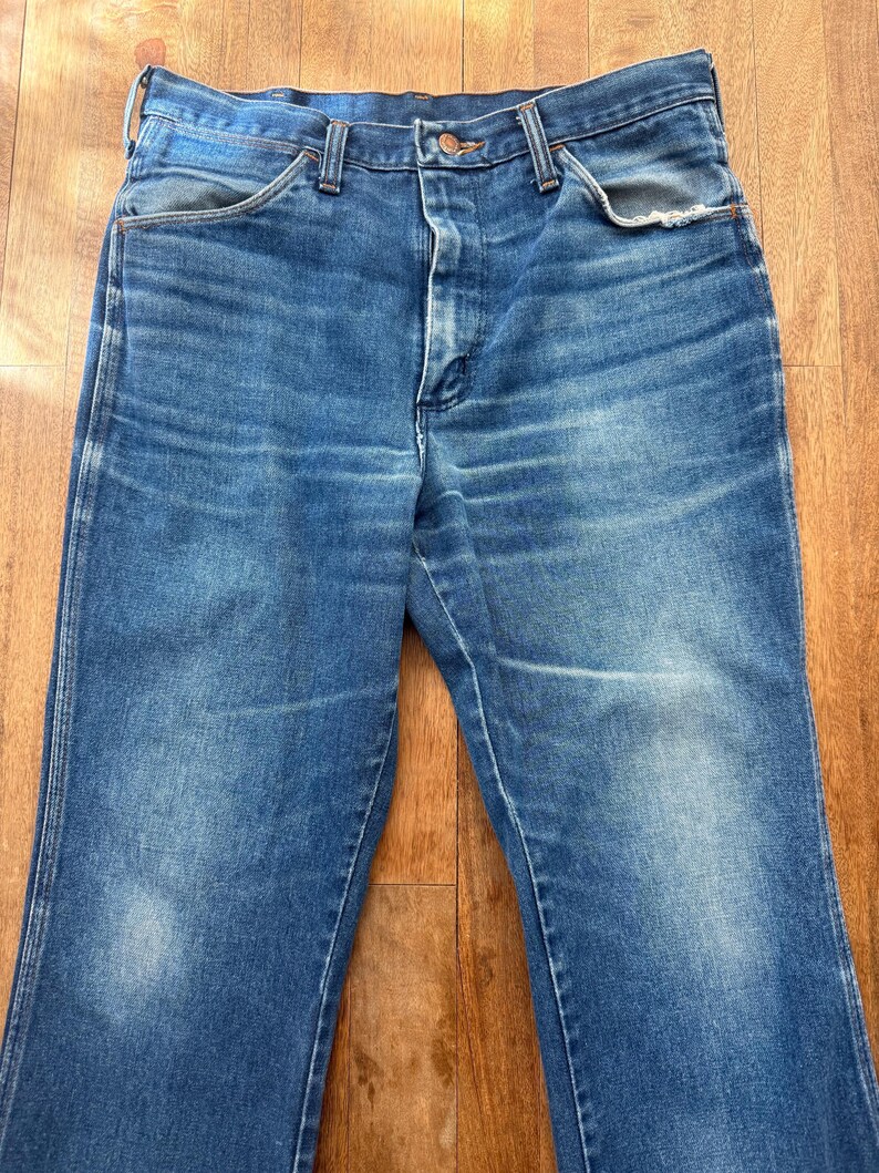 Pu&ograve; includere: Un paio di jeans vintage in denim blu. I jeans hanno un classico design a cinque tasche, una chiusura con bottone e cerniera e un lavaggio sbiadito. Il denim mostra segni di usura, con qualche sbiaditura e usura. I jeans sono appoggiati su una superficie di legno.