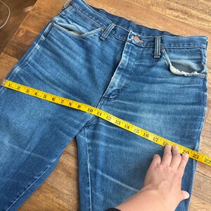 Pu&ograve; includere: Jeans blu in denim con un classico design a cinque tasche. Un metro a nastro giallo mostra una misurazione della vita di circa 51 cm. I jeans sono appoggiati su una superficie di legno, mostrando il loro aspetto sbiadito e usurato.