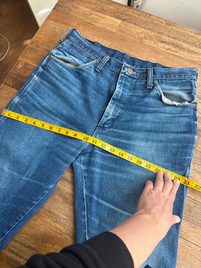 Pu&ograve; includere: Jeans blu in denim con un classico design a cinque tasche. Un metro a nastro giallo mostra una misurazione della vita di circa 51 cm. I jeans sono appoggiati su una superficie di legno, mostrando il loro aspetto sbiadito e usurato.