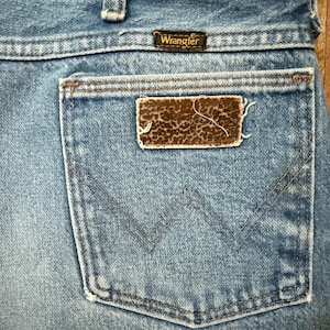May include: Close-up of the back pocket of a pair of blue denim Wrangler jeans. The pocket features a brown and tan patch with a textured pattern. The Wrangler logo is visible above the pocket.