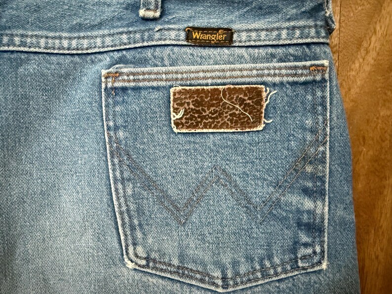 May include: Close-up of the back pocket of a pair of blue denim Wrangler jeans. The pocket features a brown and tan patch with a textured pattern. The Wrangler logo is visible above the pocket.
