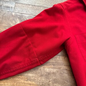 以下が含まれることがあります： 鮮やかな赤色の長袖シャツのクローズアップ。画像は袖と胴体の一部に焦点を当て、生地の質感とステッチの詳細を示しています。シャツの右側にはポケットが見えます。