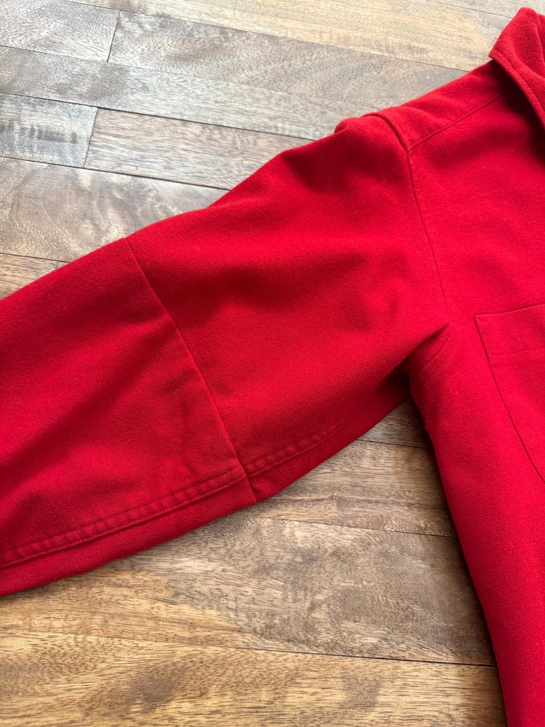 以下が含まれることがあります： 鮮やかな赤色の長袖シャツのクローズアップ。画像は袖と胴体の一部に焦点を当て、生地の質感とステッチの詳細を示しています。シャツの右側にはポケットが見えます。