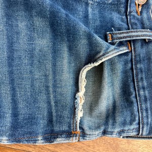 Pu&ograve; includere: Primo piano di tessuto denim blu, che mostra la trama e i dettagli di un paio di jeans. L'immagine evidenzia il bordo della tasca con dettagli sfilacciati e un passante per cintura. Il denim ha un aspetto usurato.