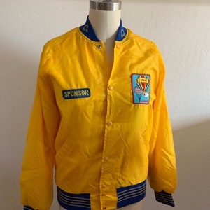 Puede incluir: Una chaqueta bomber de satén amarilla con ribetes azules y dorados. La chaqueta tiene un parche en el pecho izquierdo que dice "Sponsor" y un parche en el pecho derecho con un globo aerostático y el texto "1982 Albuquerque".