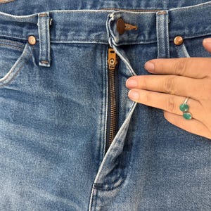 K&ouml;nnte beinhalten: Nahaufnahme einer blauen Jeans mit einem teilweise ge&ouml;ffneten Messingrei&szlig;verschluss. Die Jeans hat kupferfarbene Nieten und ein klassisches F&uuml;nf-Taschen-Design. Der Stoff zeigt eine verblasste Waschung, typisch f&uuml;r getragenen Denim.