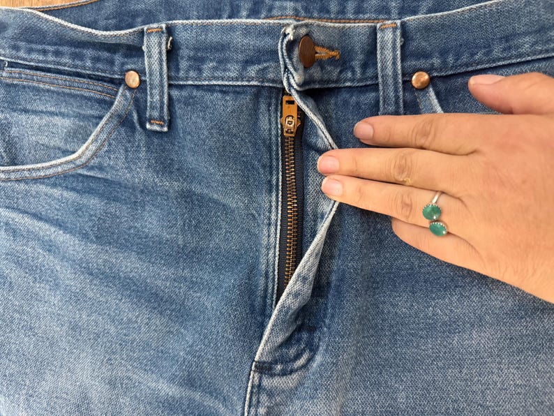 K&ouml;nnte beinhalten: Nahaufnahme einer blauen Jeans mit einem teilweise ge&ouml;ffneten Messingrei&szlig;verschluss. Die Jeans hat kupferfarbene Nieten und ein klassisches F&uuml;nf-Taschen-Design. Der Stoff zeigt eine verblasste Waschung, typisch f&uuml;r getragenen Denim.