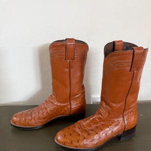 May include: A pair of brown leather cowboy boots with a textured ostrich leg design. The boots have a pointed toe, a stacked heel, and a pull-on loop. The boots are set on a dark green surface.