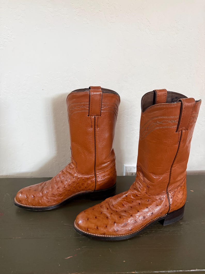 May include: A pair of brown leather cowboy boots with a textured ostrich leg design. The boots have a pointed toe, a stacked heel, and a pull-on loop. The boots are set on a dark green surface.
