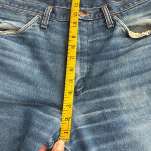 Pu&ograve; includere: Primo piano di jeans blu con un metro a nastro giallo. Il nastro mostra una misura di circa 38 cm. I jeans hanno un classico stile a cinque tasche e una chiusura con bottone e cerniera.