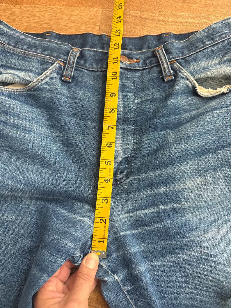 Pu&ograve; includere: Primo piano di jeans blu con un metro a nastro giallo. Il nastro mostra una misura di circa 38 cm. I jeans hanno un classico stile a cinque tasche e una chiusura con bottone e cerniera.