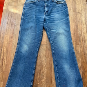 Pu&ograve; includere: Jeans blu in denim con gamba a zampa. I jeans presentano un classico design a cinque tasche, una chiusura con bottone e cerniera e mostrano segni di usura. Il denim ha un aspetto sbiadito, con aree pi&ugrave; chiare sulle cosce.