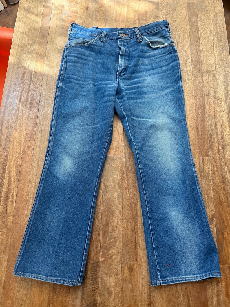 Pu&ograve; includere: Jeans blu in denim con gamba a zampa. I jeans presentano un classico design a cinque tasche, una chiusura con bottone e cerniera e mostrano segni di usura. Il denim ha un aspetto sbiadito, con aree pi&ugrave; chiare sulle cosce.