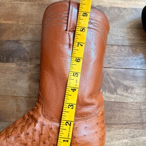 May include: A brown leather cowboy boot with a textured surface, measured with a yellow tape measure. The boot is approximately 10 inches tall. The boot has a pointed toe and a stacked heel.
