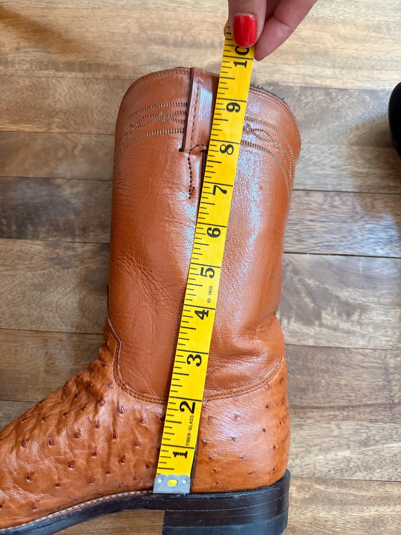 May include: A brown leather cowboy boot with a textured surface, measured with a yellow tape measure. The boot is approximately 10 inches tall. The boot has a pointed toe and a stacked heel.