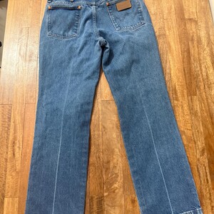 May include: Light blue denim jeans with a classic five-pocket design. The jeans feature a brown leather patch on the back pocket and a straight leg cut. The jeans are laid flat on a wooden surface.
