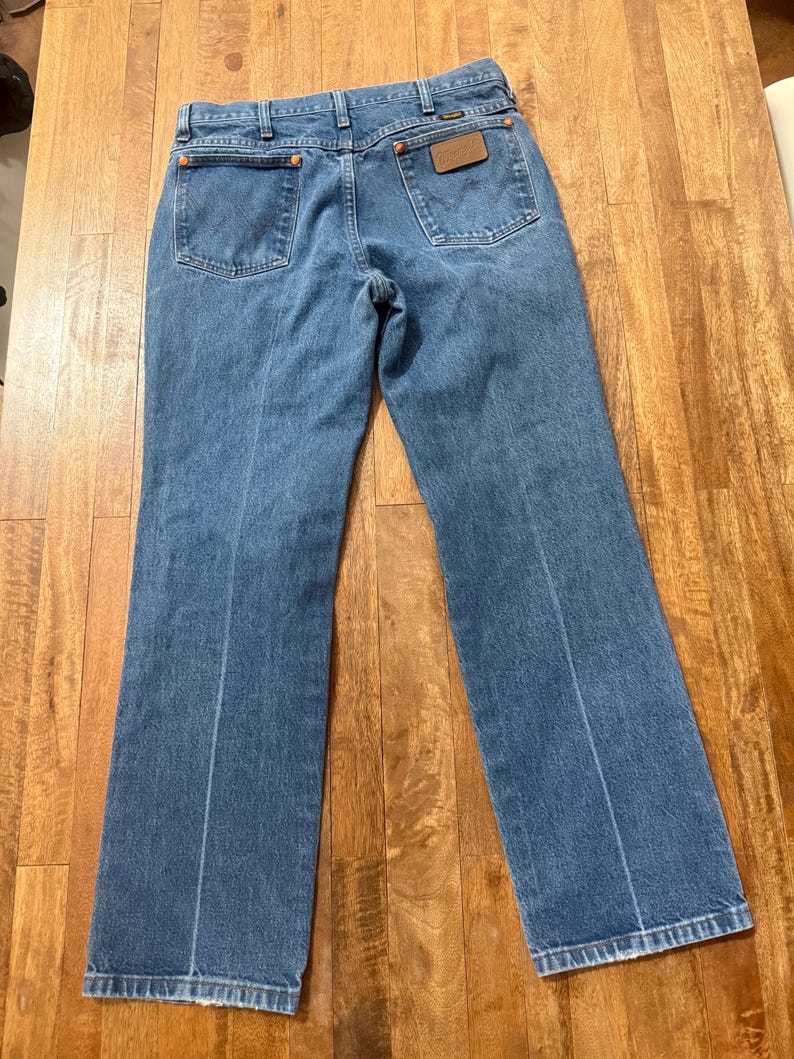 May include: Light blue denim jeans with a classic five-pocket design. The jeans feature a brown leather patch on the back pocket and a straight leg cut. The jeans are laid flat on a wooden surface.