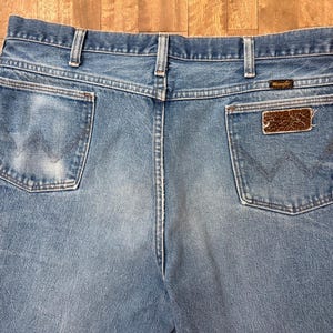 May include: Light blue denim jeans with the Wrangler logo on the back pocket. The jeans have a classic five-pocket design and a faded wash. The back pockets feature the iconic Wrangler stitching.