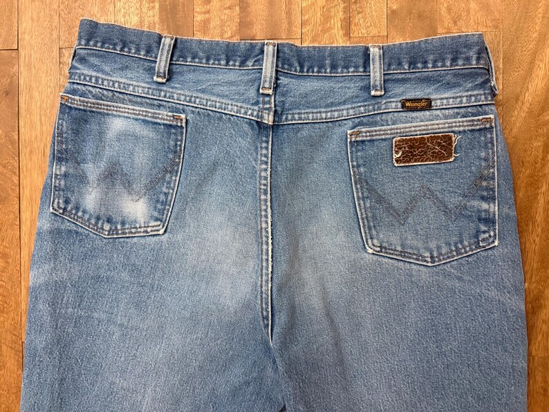 May include: Light blue denim jeans with the Wrangler logo on the back pocket. The jeans have a classic five-pocket design and a faded wash. The back pockets feature the iconic Wrangler stitching.